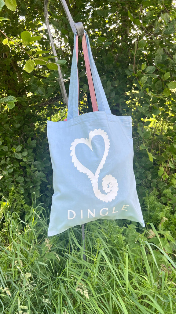 Light blue tote bag with a white heart design and 'Dingle' text, hanging on a tree branch with green foliage in the background.